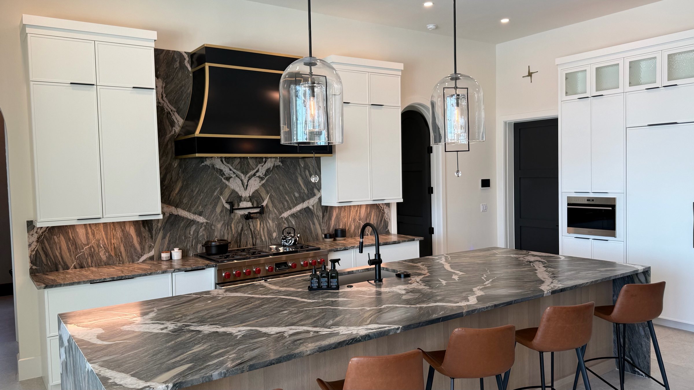 Veined stone island and full-height backsplash under a black-and-brass hood